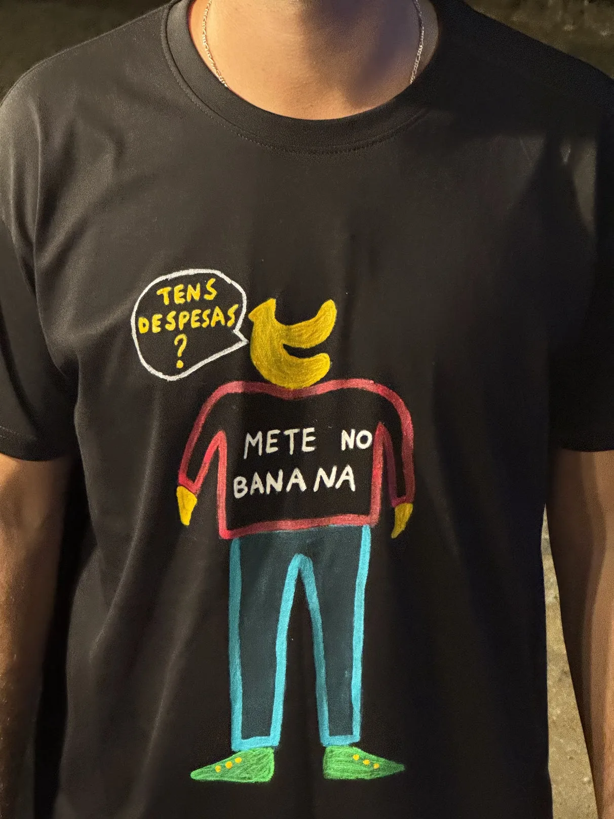 Black hand-painted t-shirt with a banana character and the Portuguese phrases Tens despesas? and Mete no Banana