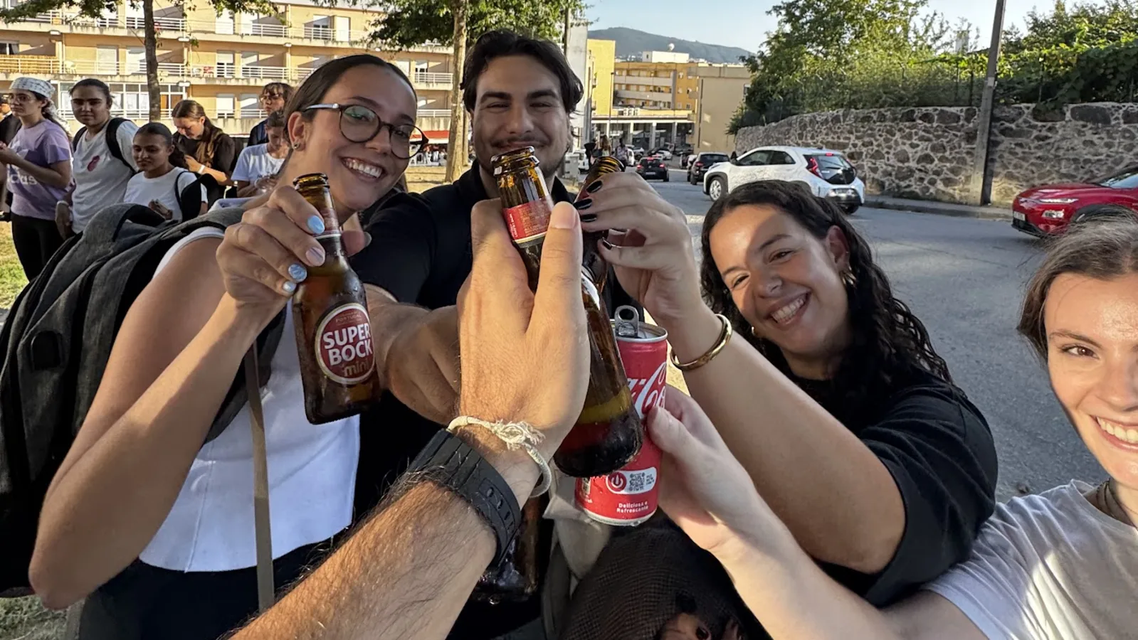 Four people raising beer bottles and a can in a toast in the late afternoon, next to the UMinho campus