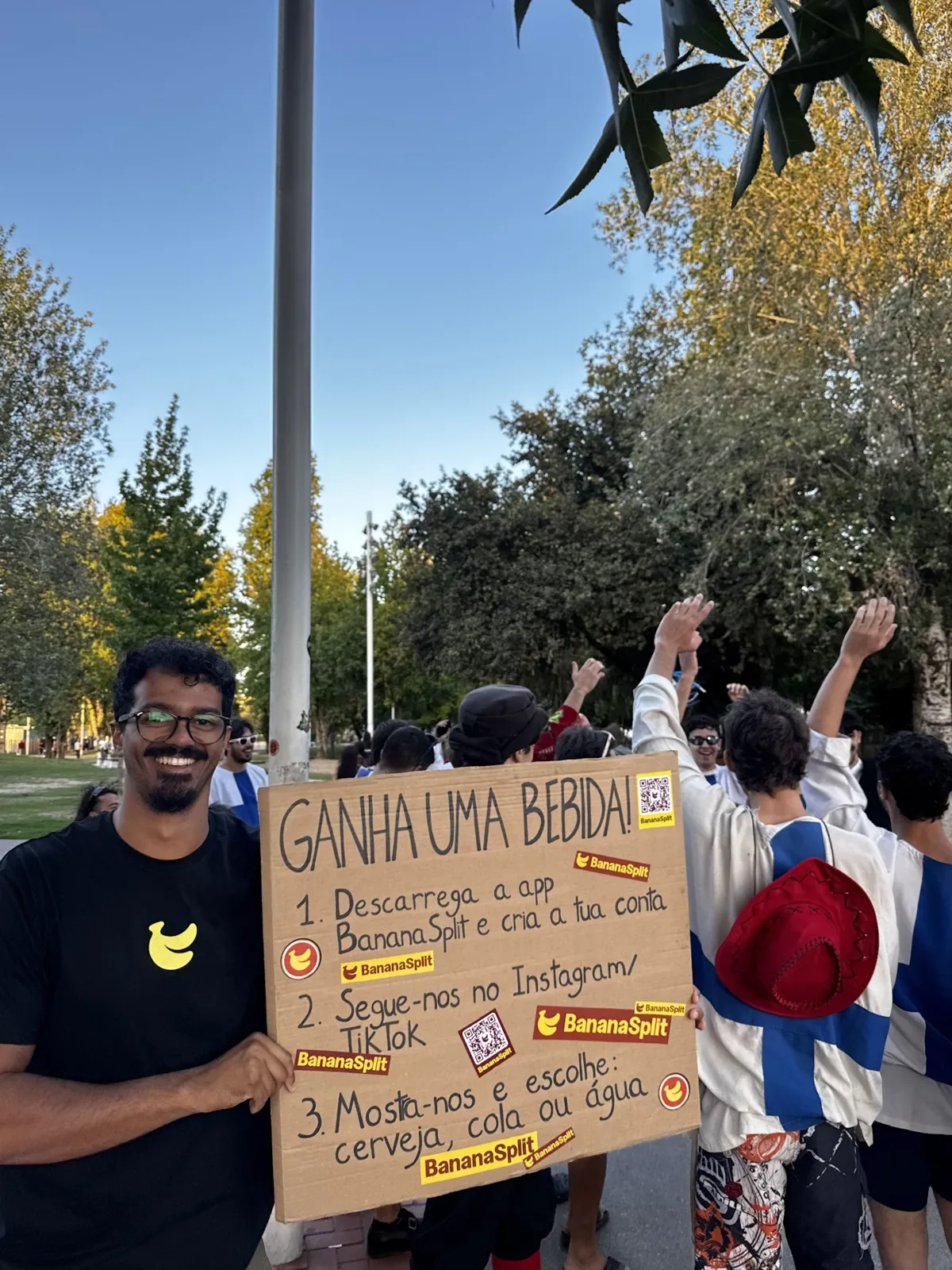 Leo holding the "Win a drink!" sign in the Rodovia park, with a group of students celebrating behind him